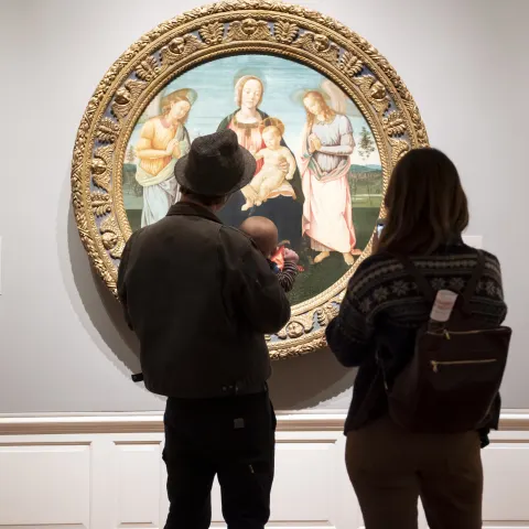 two people and a baby stand in front of a round oil classical painting of a religious scene at the Portland Art Museum